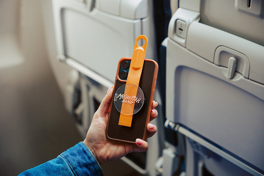 Traveler holding an Orange Mo'Bettah Airplane Phone Holder next to a MagSafe-compatible iPhone